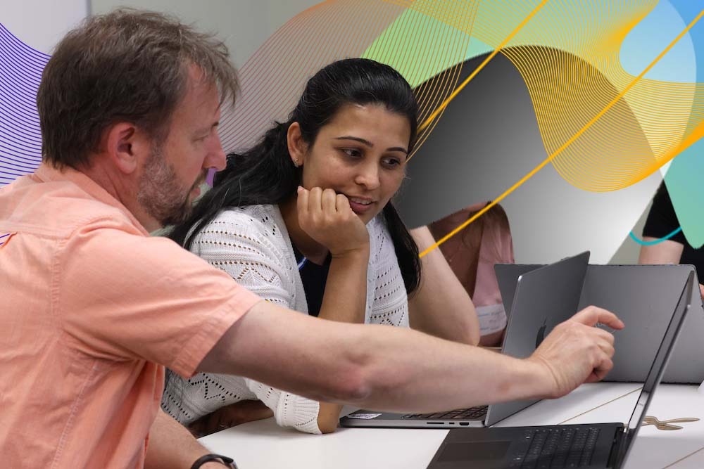 Two people sitting at a table collaborating on a laptop, with one person pointing at the screen while the other looks on attentively.